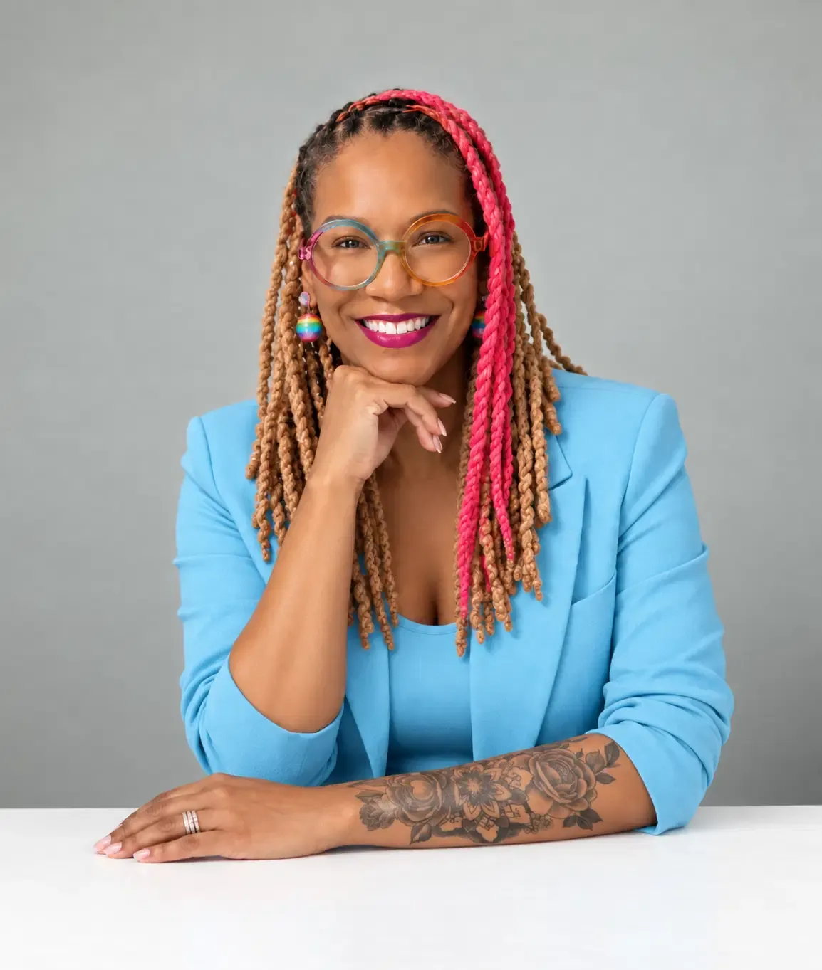 Angela Briggs-Paige smiling in a light blue suit at a white table with her hand under her chin against a grey studio background