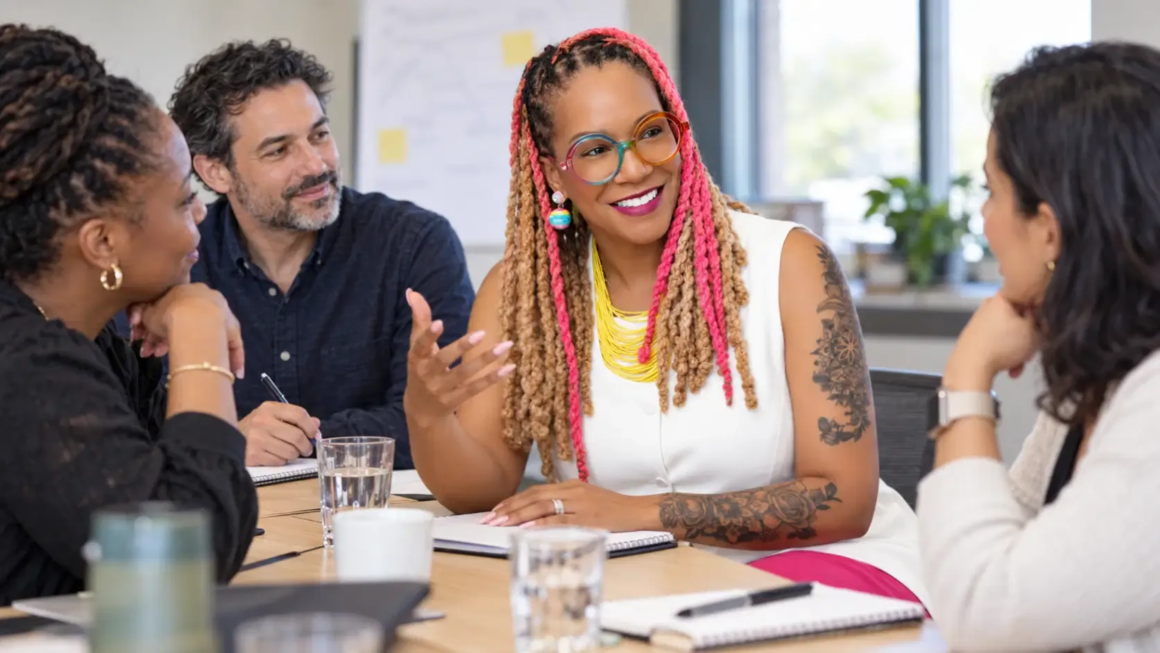 Angela Briggs-Paige in a white sleeveless top with bright pink-and-blonde braids and colorful glasses, seated at a light-wood workshop table with four colleagues